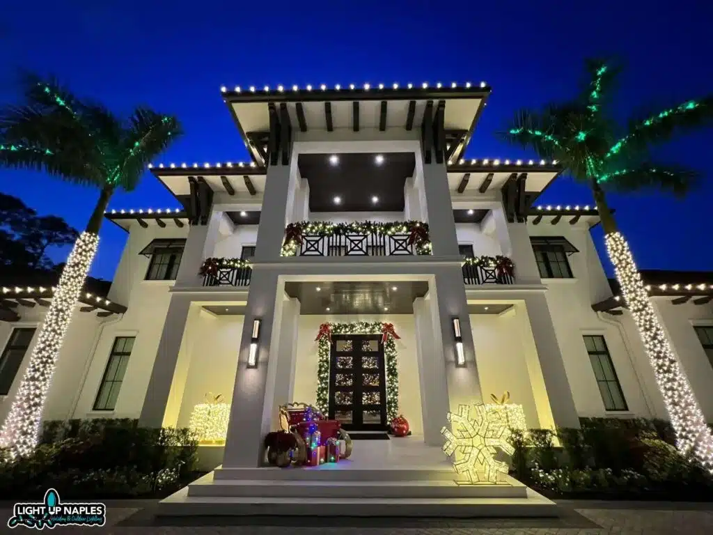 Light Up Naples full Christmas light installation with roofline lights, green palm tree wrapping, balcony garland, door surround, and lighted gift displays on a FL estate